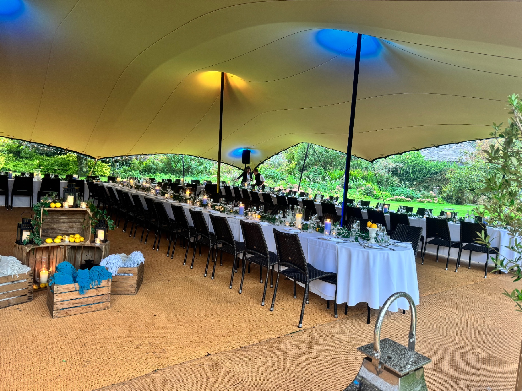 Long banqueting tables under a stretch tent in an English garden, dressed with candles, lemons and eucalyptus for a summer dinner