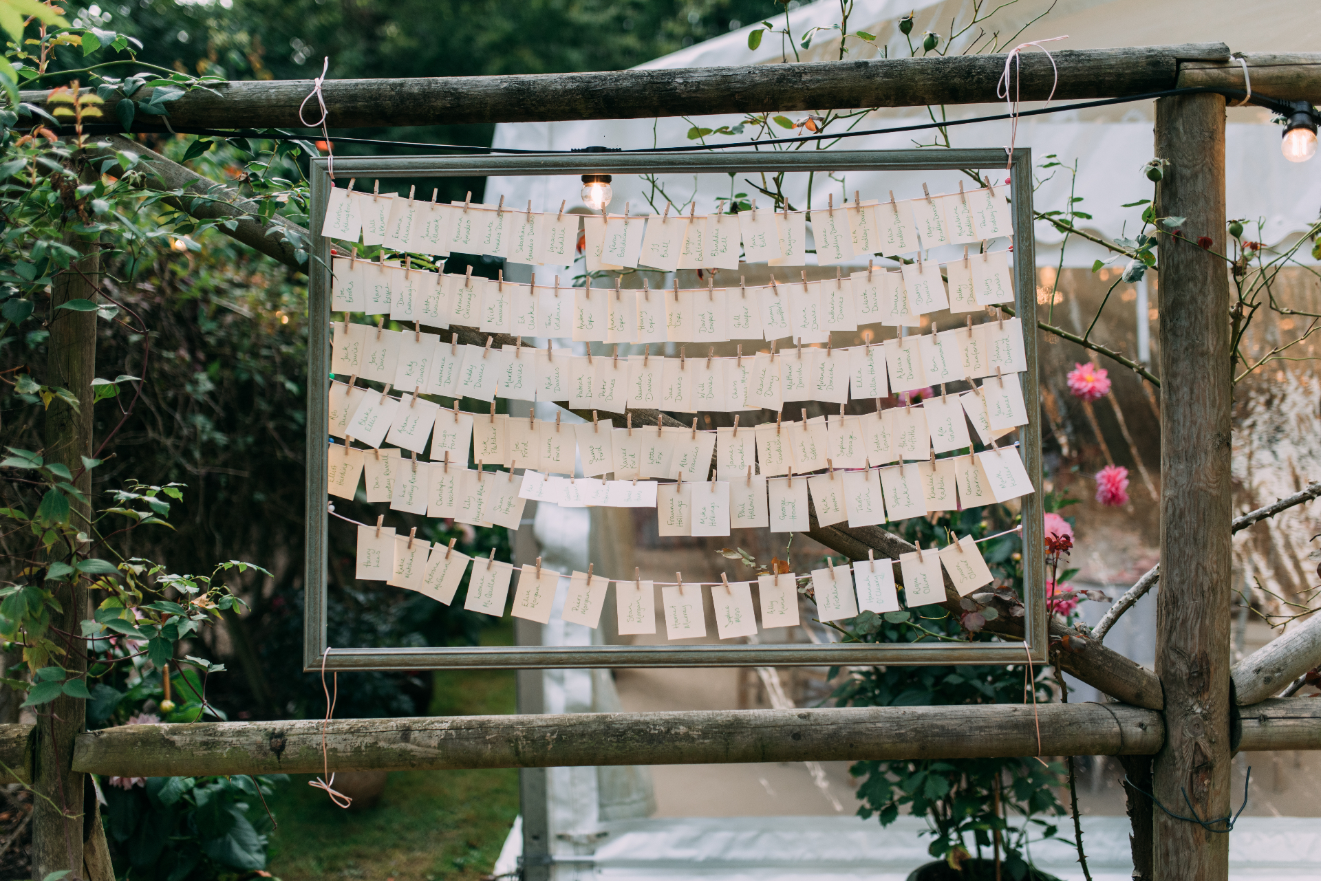 Wedding seating chart with guest name cards displayed on strings in a rustic wooden frame surrounded by greenery