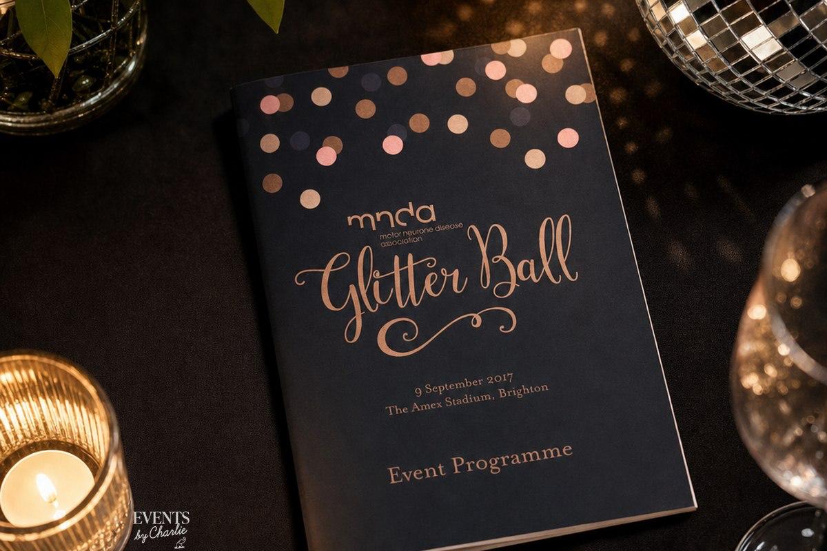 MNDA Glitter Ball event programme in rose gold script on a navy cover, styled with candlelight and a mirrored disco ball