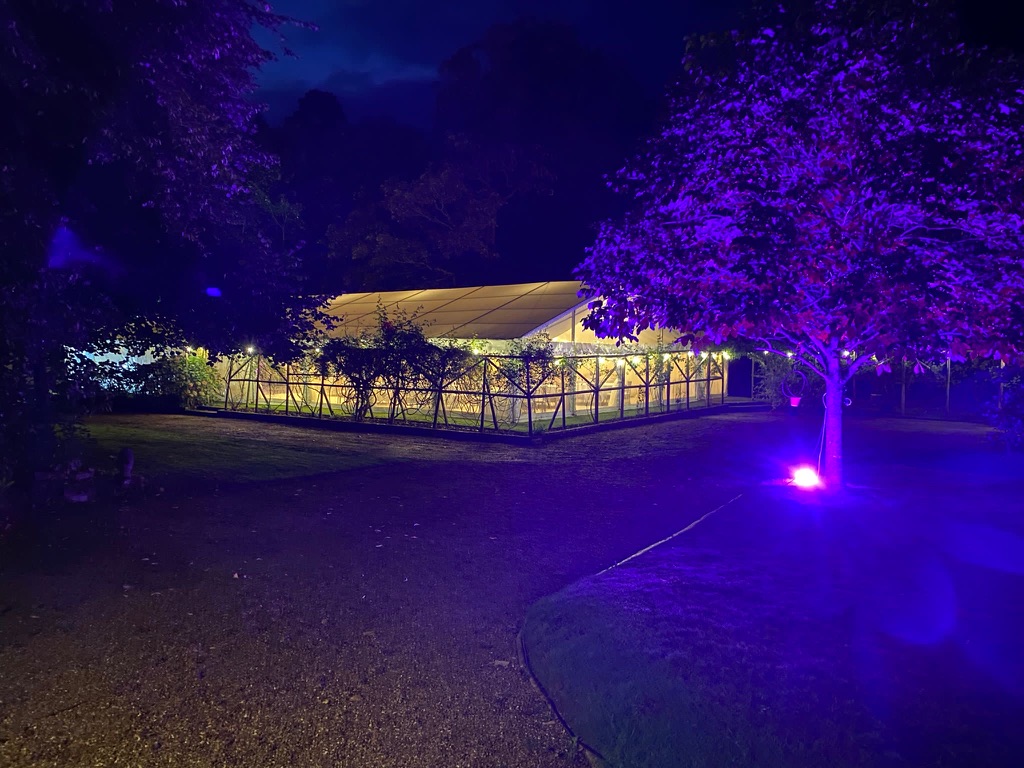 Marquee glowing warmly at night with a tree lit in purple in the garden