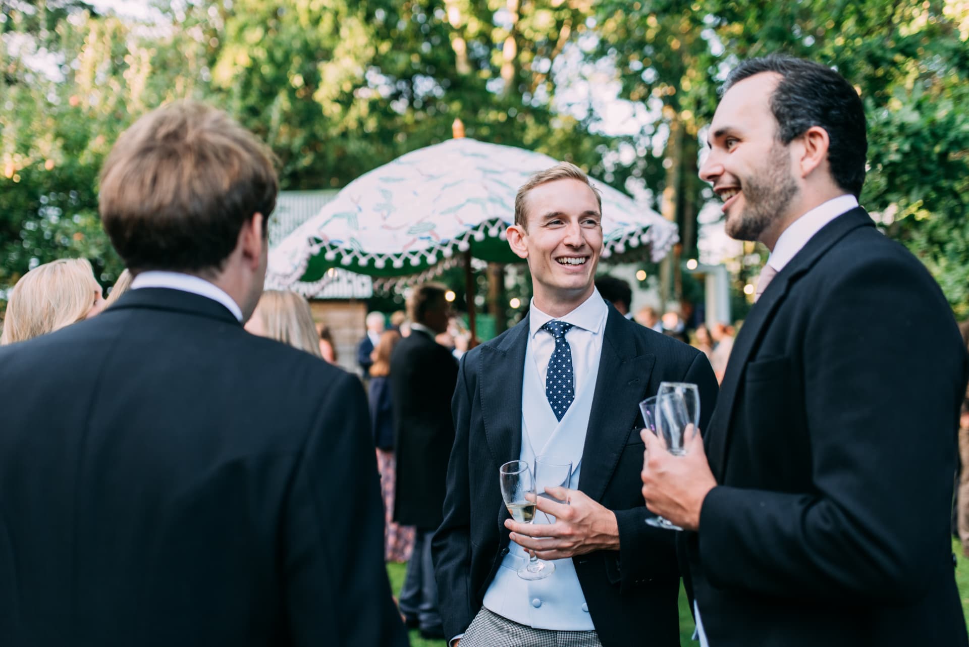 Wedding guests in morning suits laughing over champagne at a garden drinks reception