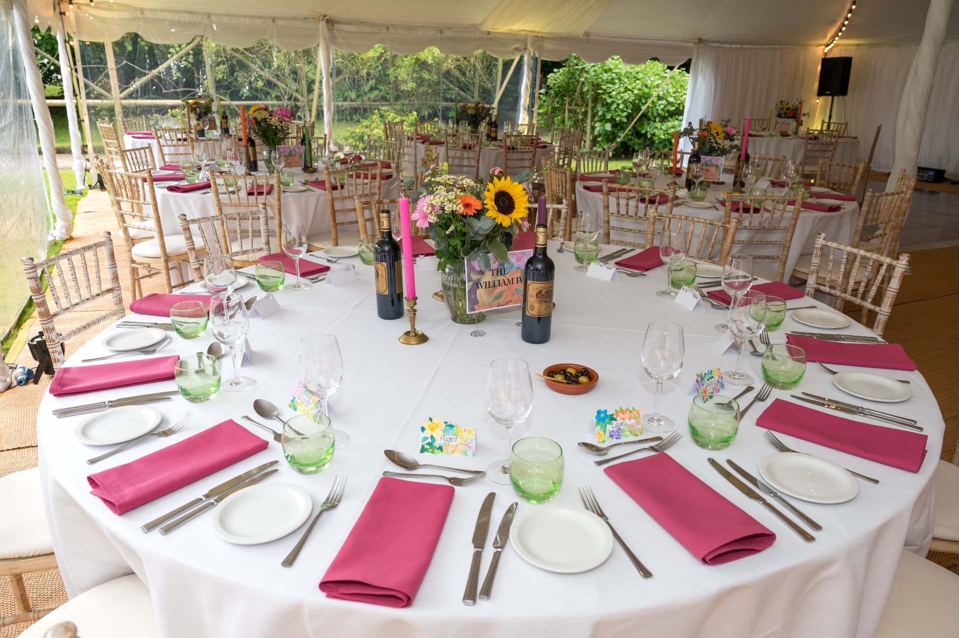 Round table laid with white linen, pink napkins and a sunflower centrepiece in a clear-sided marquee