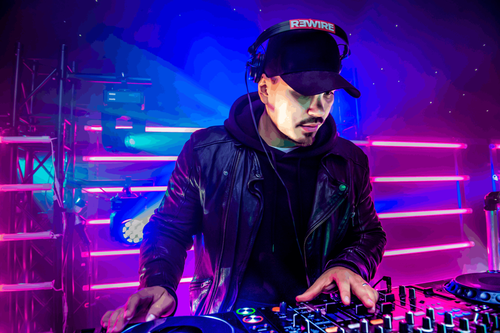 DJ at Pioneer decks wearing a REWIRE cap, lit by pink and blue neon strip lighting