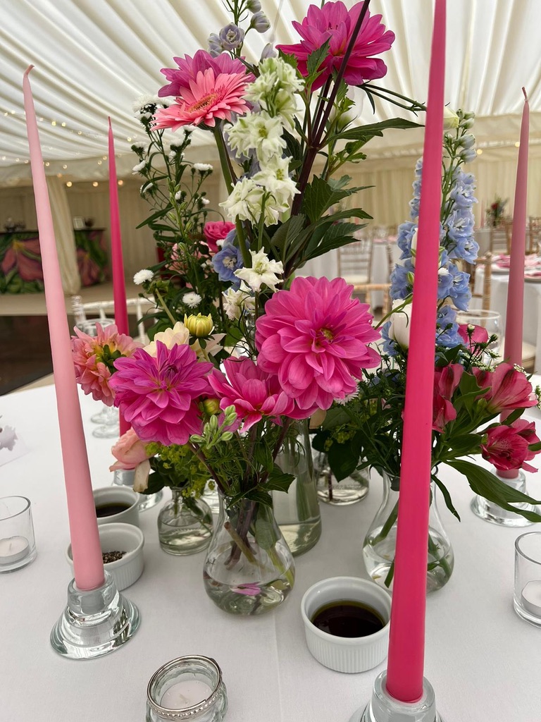 Bud vases of pink dahlias, gerbera and delphinium with tall pink tapered candles on a wedding table