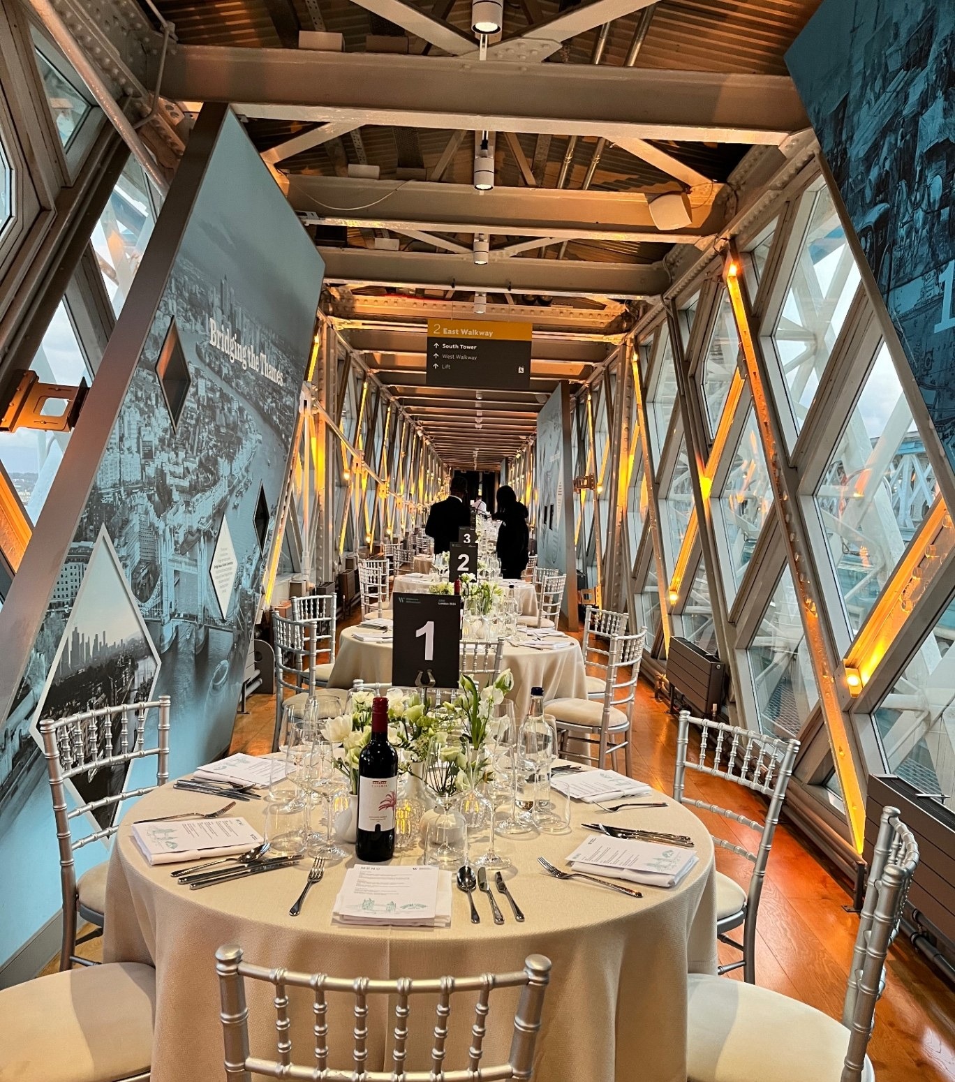Elegant round tables dressed in ivory linen with silver chiavari chairs, laid up for a dinner inside Tower Bridge's glass walkway
